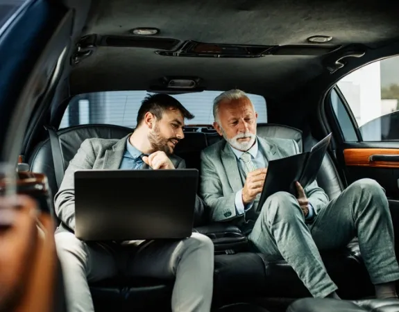 Two businessmen are working with laptops inside a luxury limousine.