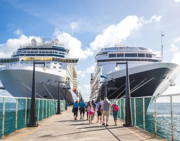 Two large cruise ships docked at a pier with people walking between them under a clear blue sky.