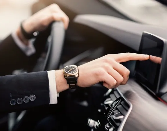 An expert chauffeur driving a limo, pointing at the touchscreen dashboard while wearing a wristwatch.