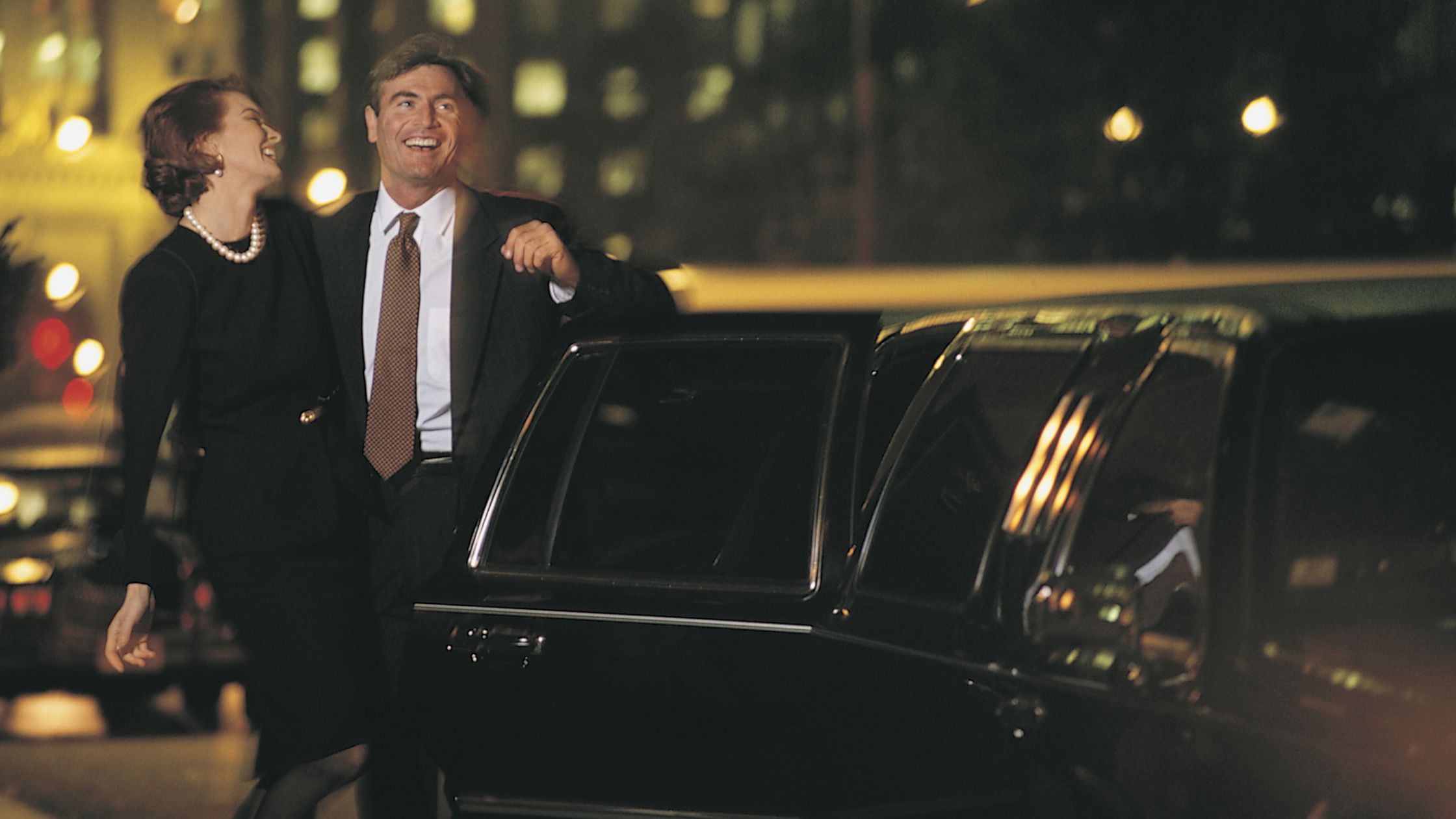 Smiling couple in formal attire beside black limousine at night