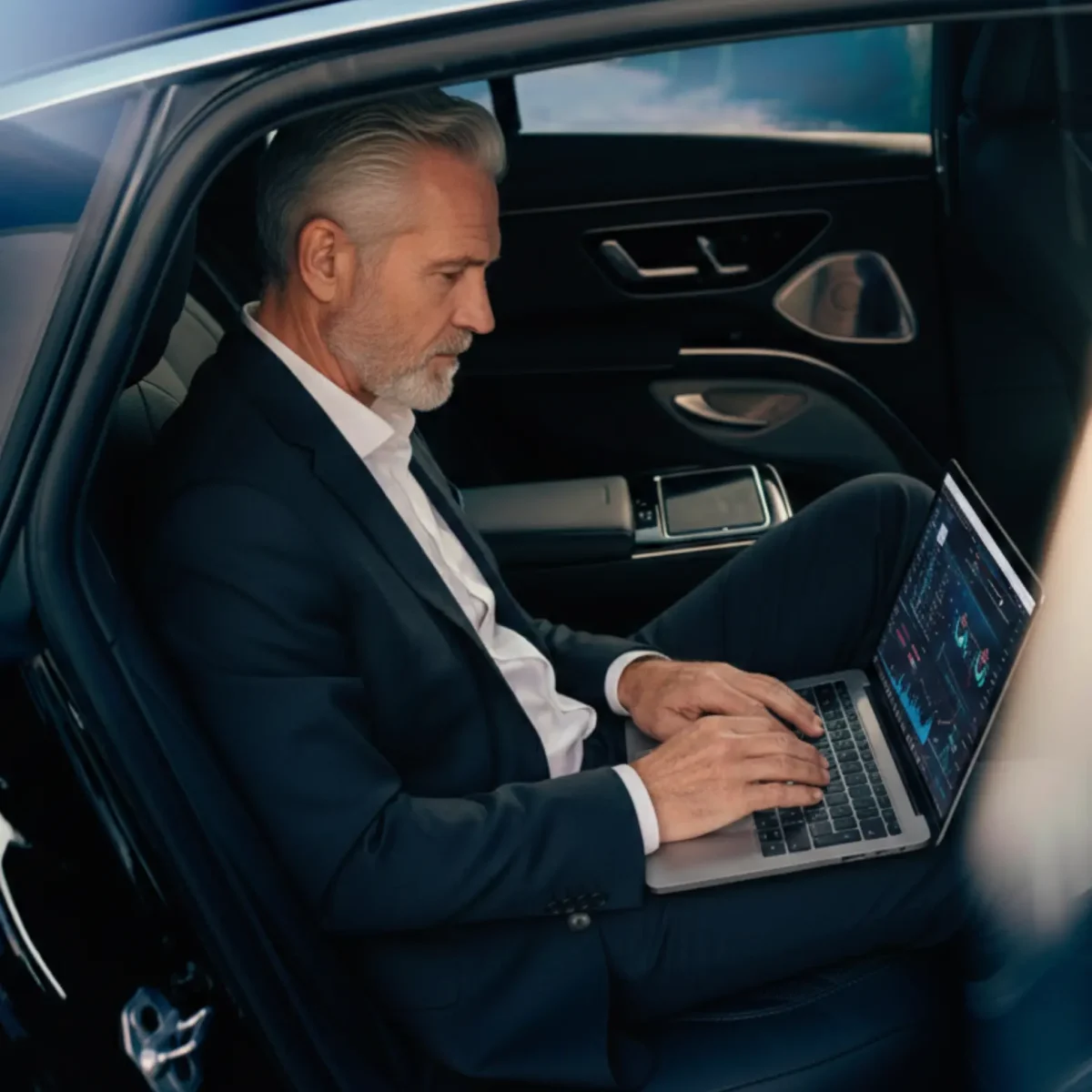 A businessman in a suit working on a laptop while sitting in the back seat of a premium executive sedan