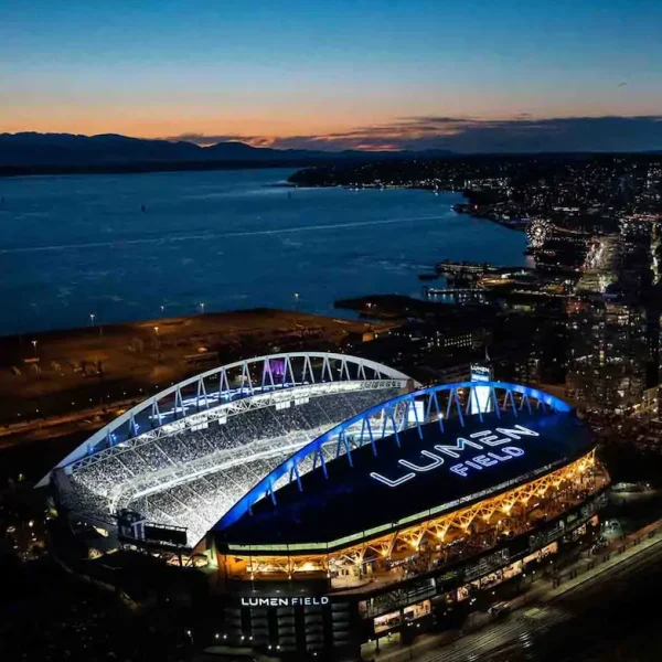Aerial view of Lumen Field stadium at night lit up in blue next to the Seattle waterfront and Elliott Bay
