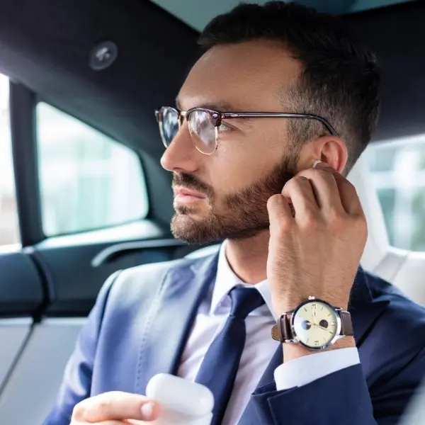 Close-up of a bearded man in a professional blue suit and tie sitting in a luxury car's backseat. He is wearing glasses and a classic wristwatch while adjusting a white wireless earbud in his ear and looking thoughtfully out the window.